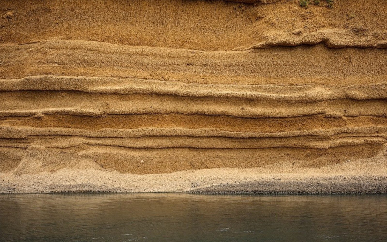 A raw eroded riverbank several metres high showing horizontal layers of sand and silt, water at its foot